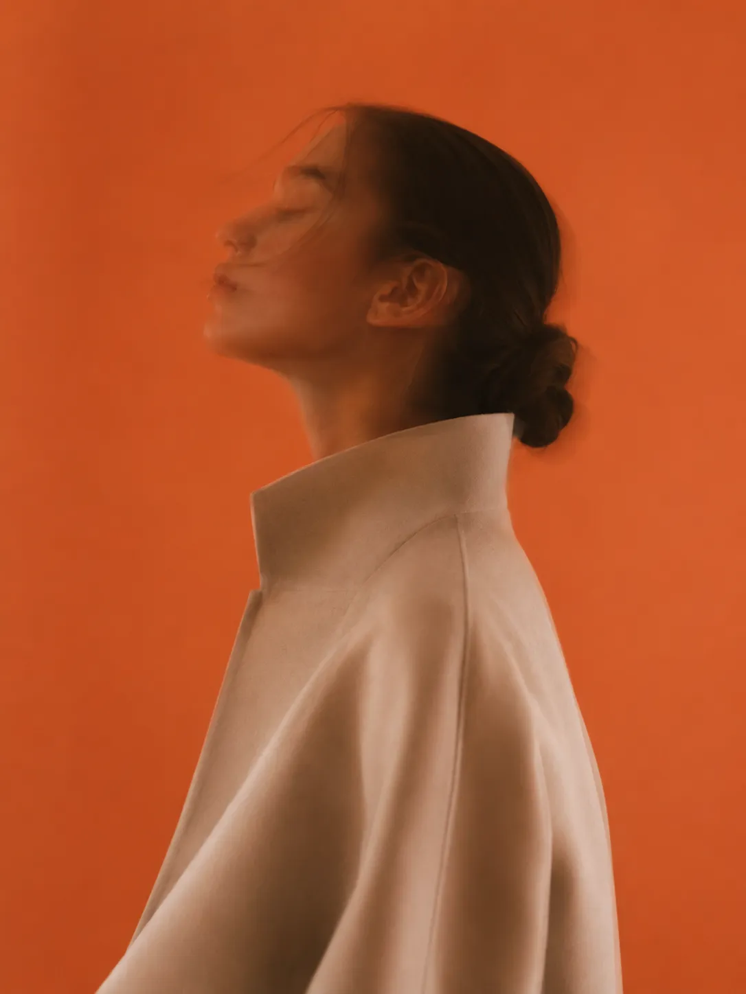 A side profile of a woman with her hair in a low bun and eyes closed, wearing a high-collared cream coat against a warm orange background.