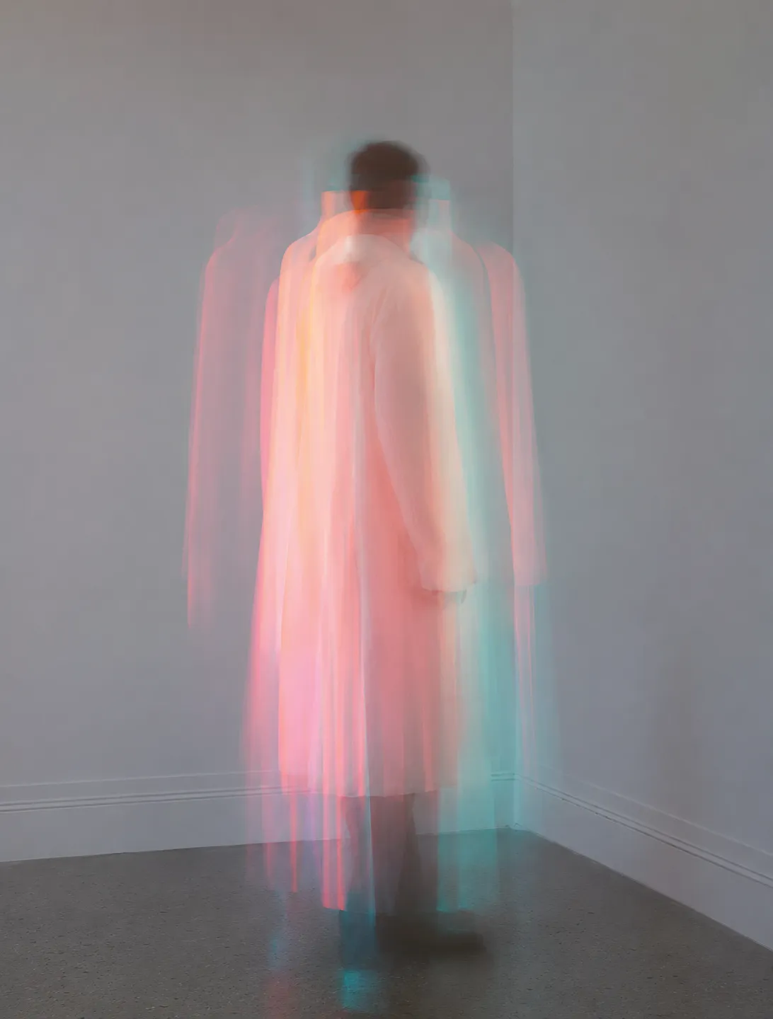 A long-exposure photograph of a person standing in a bare room, their figure blurred into overlapping pink and teal trails of movement.