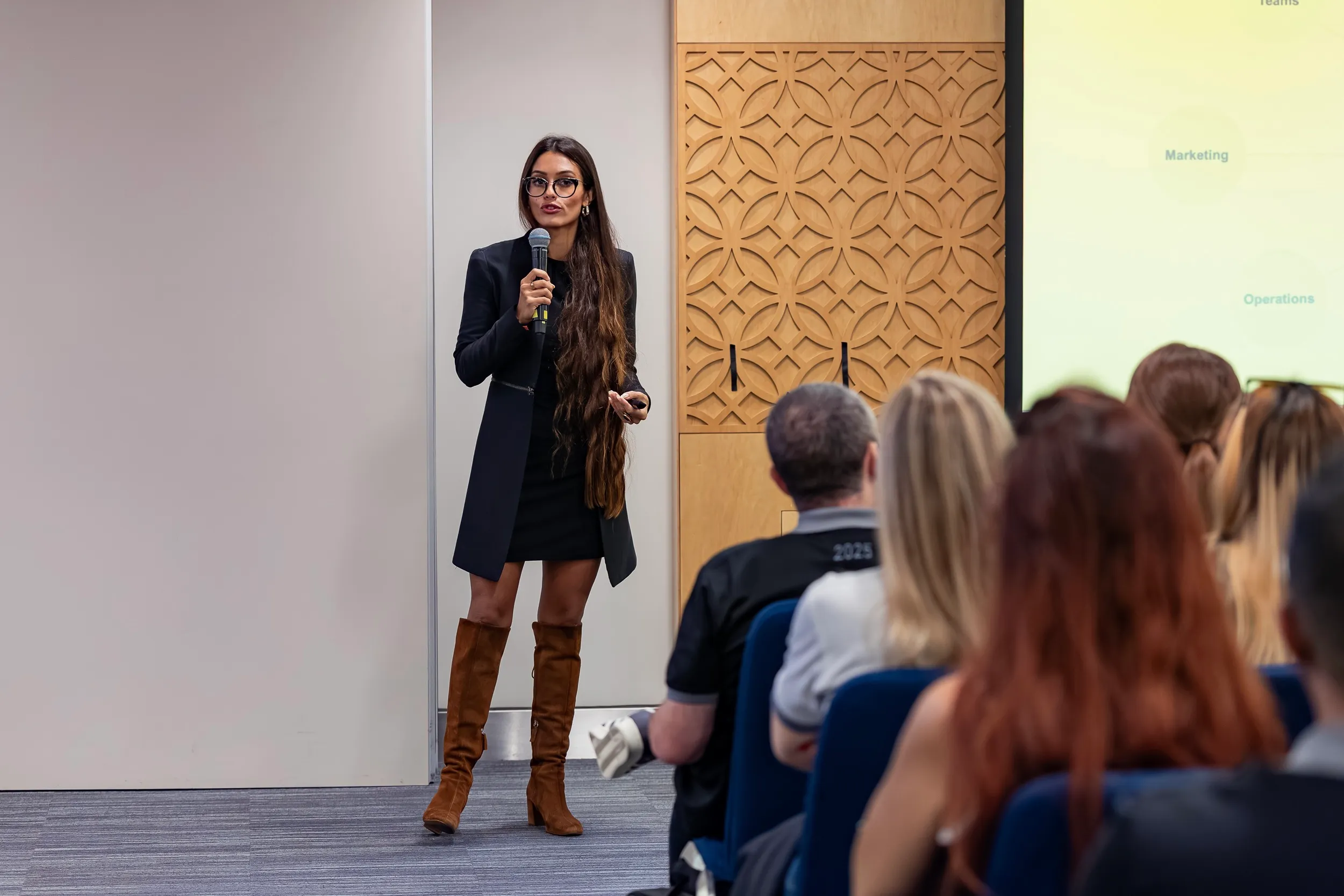Ivy Forero speaking with a microphone to a seated audience, a presentation slide projected behind her